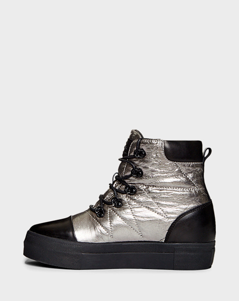 The Rudsak TASSEL - CA EN 8219514 SILVER is a stylish high-top sneaker with a metallic silver quilted body, black toe cap, heel, and sole, black laces with metal eyelets, and faux shearling lining. Shown on a plain light gray background.