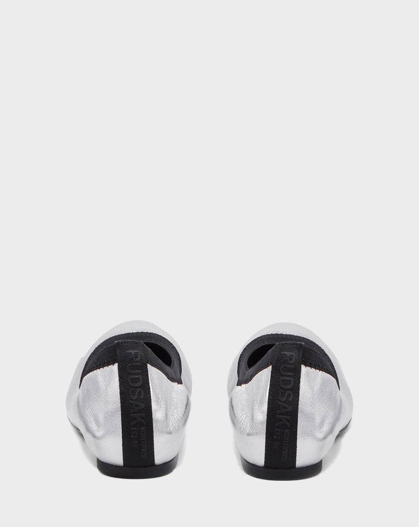 The TESSALY - CA EN 8220037 SILVER by Rudsak are silver slip-on shoes with black pull tabs featuring the vertical Rudsak branding, combining ballerina leather elegance with a comfy wedge for all-day wear.