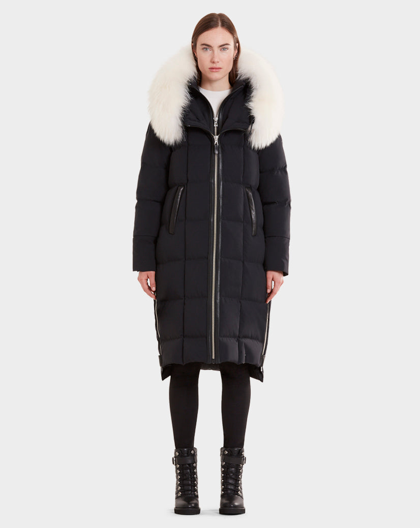 A woman stands facing forward wearing the Rudsak MILANI - CA EN 8119582B BLACK OFF WHITE goose down puffer coat with an oversized white fur hood, paired with black leggings and lace-up boots against a plain light background.