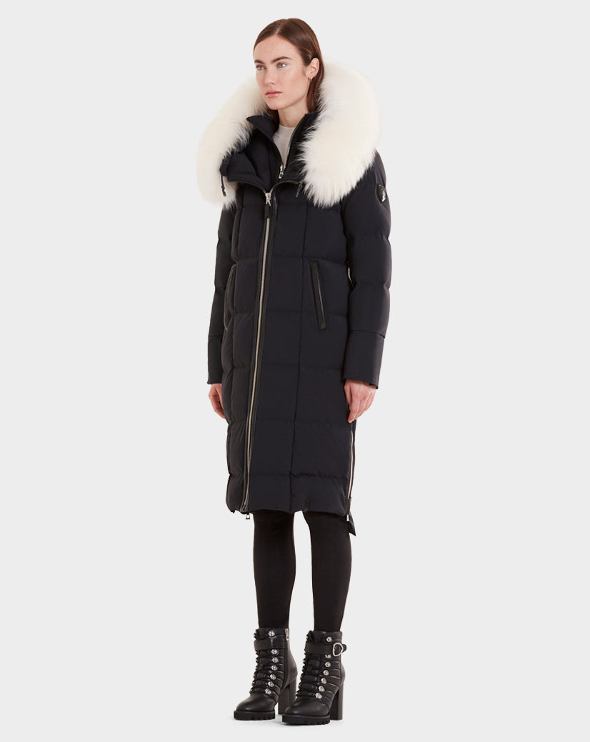 A woman wears the Rudsak MILANI - CA EN 8119582B BLACK OFF WHITE, a long black goose down puffer with an oversized white fur-trimmed hood, paired with black leggings and heeled lace-up boots against a plain light background.