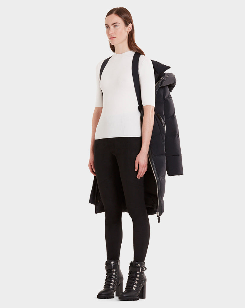 A woman with straight brown hair and a neutral expression faces forward, wearing the Rudsak MILANI - CA EN 8119582B BLACK OFF WHITE fitted top and black quilted parka draped over her shoulders, styled with black leggings and lace-up heeled boots.