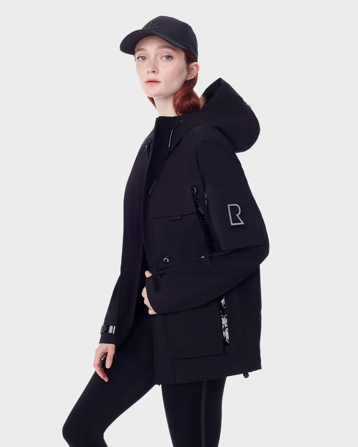 A woman models the RUDSAK PALM - 9123000 BLACK F recycled Stormshell rainwear jacket with large pockets and sleeve logo, paired with black leggings and a matching cap, against a plain light gray background.