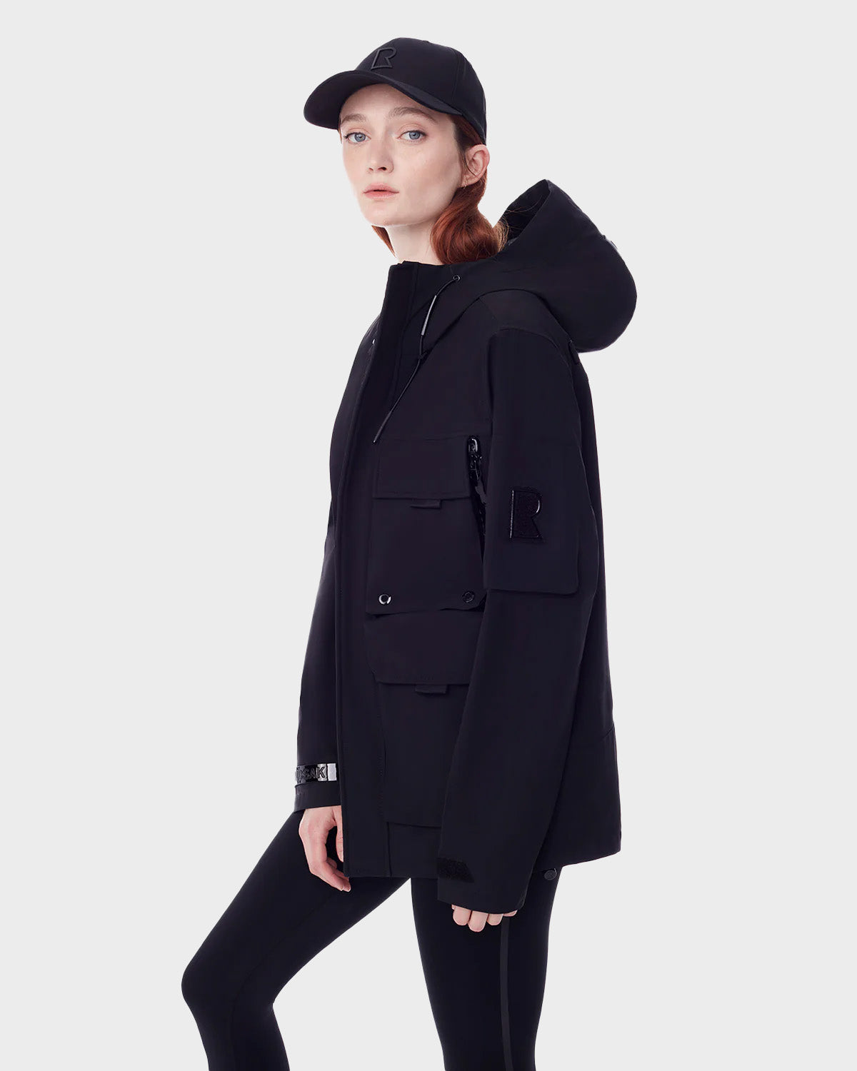 A woman with fair skin and red hair wears the RUDSAK PALM - 9123000 BLACK F utility rainwear jacket with black leggings and a black cap, standing sideways against a plain light background.
