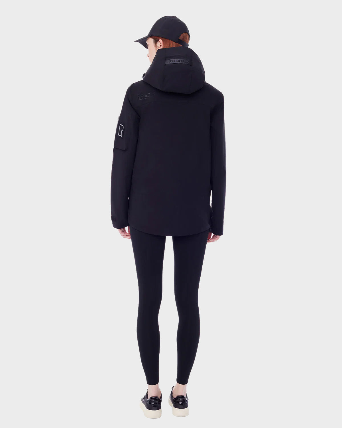 A person stands facing away against a light background, wearing the RUDSAK PALM - 9123000 BLACK F utility rainwear jacket, paired with black leggings, a black cap, and black slip-on shoes with white soles.