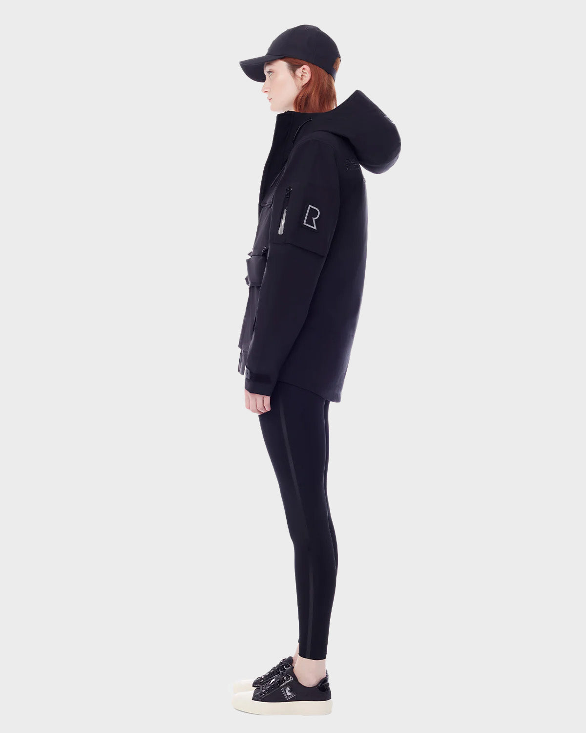 A person in profile wears the RUDSAK PALM - 9123000 BLACK F unisex rain jacket with black leggings, a black cap, and black sneakers with white soles, standing against a plain light gray background.