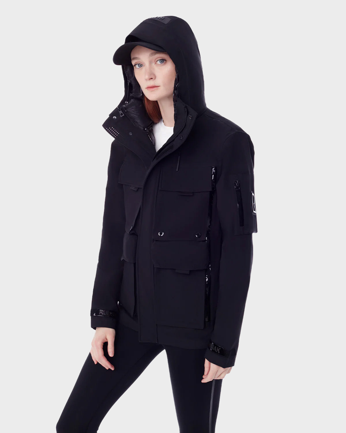 A woman in the RUDSAK PALM - 9123000 BLACK F utility rainwear jacket, black cap, and leggings stands against a light gray background, gazing to the side with a neutral expression.
