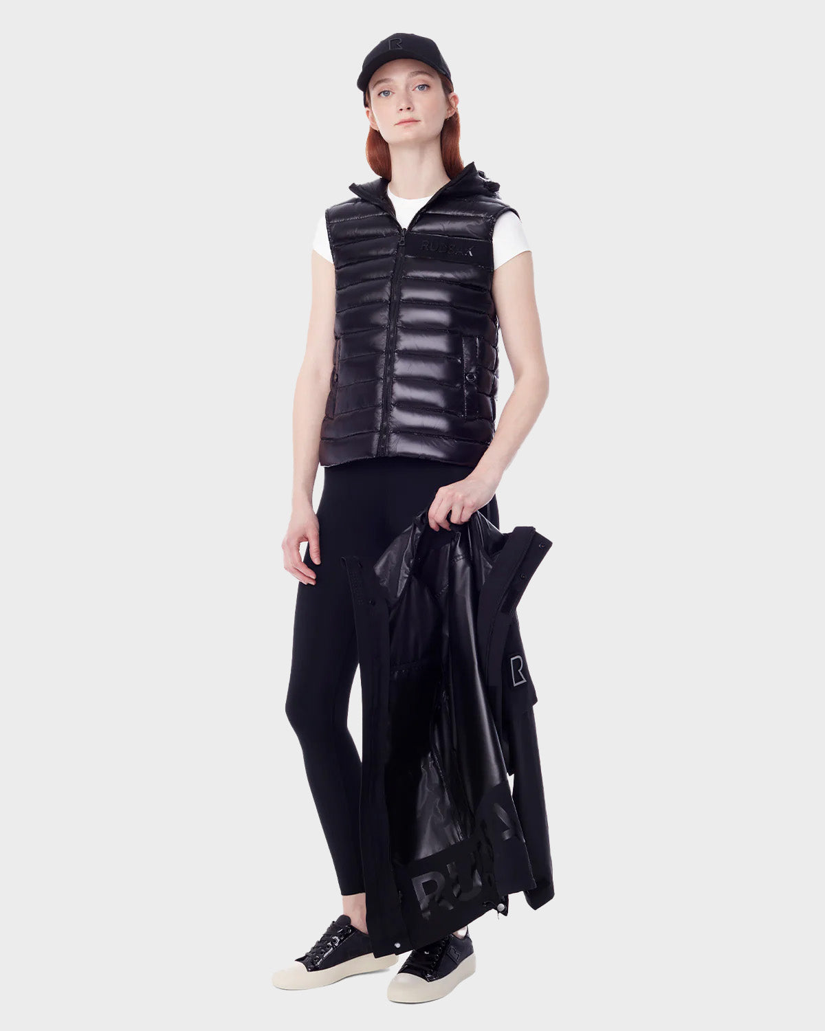 A person holds the RUDSAK PALM - 9123000 BLACK F recycled Stormshell utility rainwear jacket, wearing a black cap, black puffer vest, white t-shirt, black leggings, and black sneakers against a light gray background.