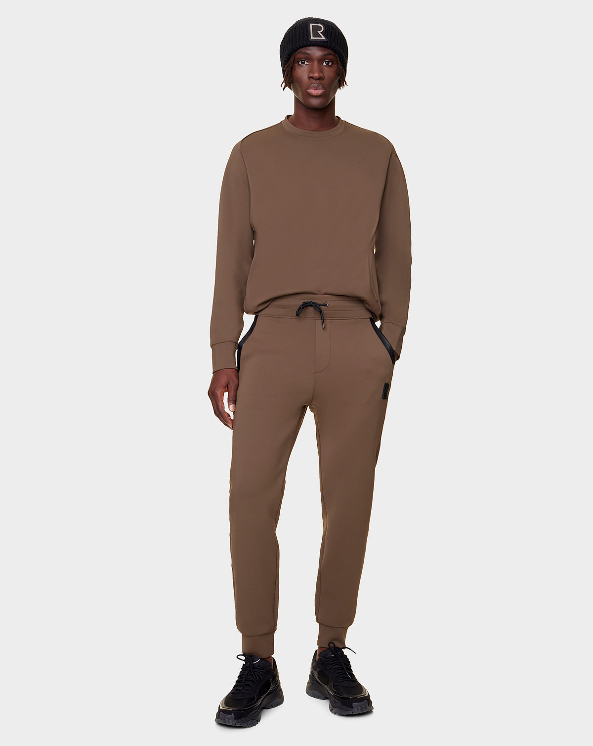 A person stands facing forward in Rudsak's OLAV - 9522503 ARMY MEN matching brown sweatshirt and unisex joggers, plus a black beanie with a B logo and black sneakers, against a plain light background.