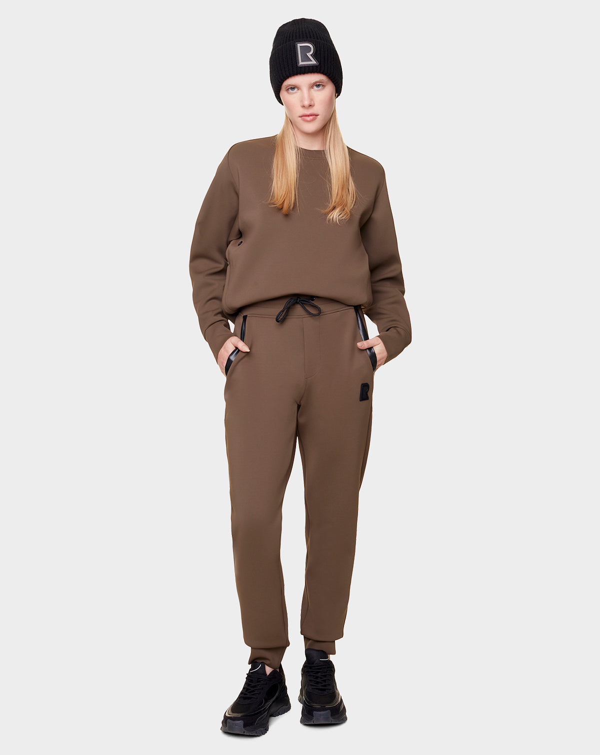 A woman faces forward in Rudsak’s OLAV - 9522503 ARMY WOMEN joggers with a matching brown sweatshirt, black sneakers, and a black beanie with a white “R.” She has long blonde hair and her hands are in her pockets.