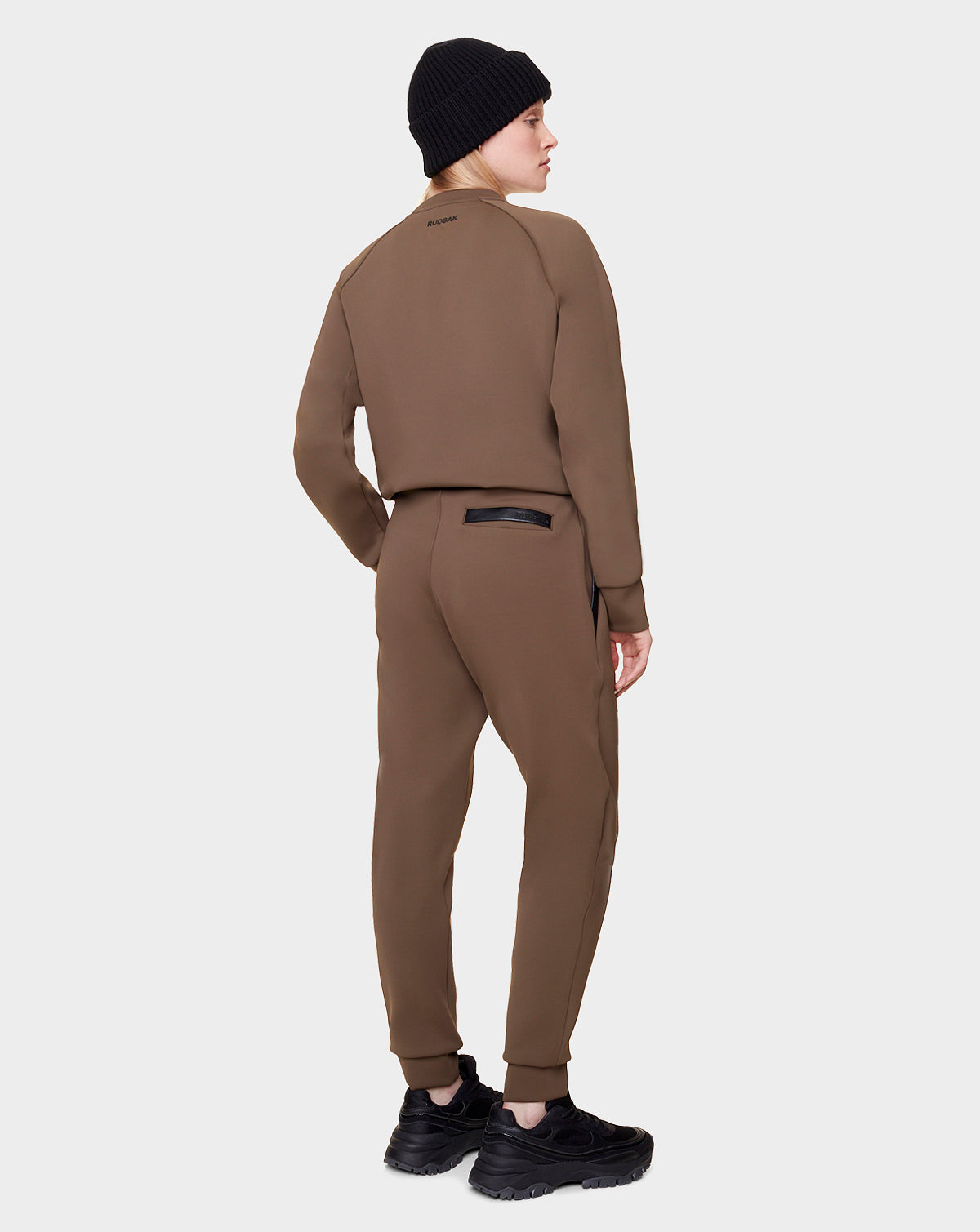 A person in an army green Rudsak OLAV - 9522503 women's tracksuit, with a black beanie and sneakers, stands facing away and looking to the side against a plain light background.