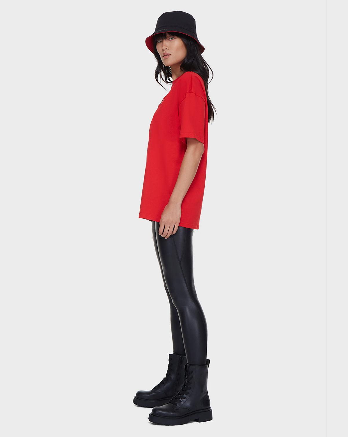 A woman stands sideways against a plain light gray background, wearing a black bucket hat, Rudsak LARY - 9521501 POPPY RED unisex t-shirt, black shiny leggings, and black combat boots.