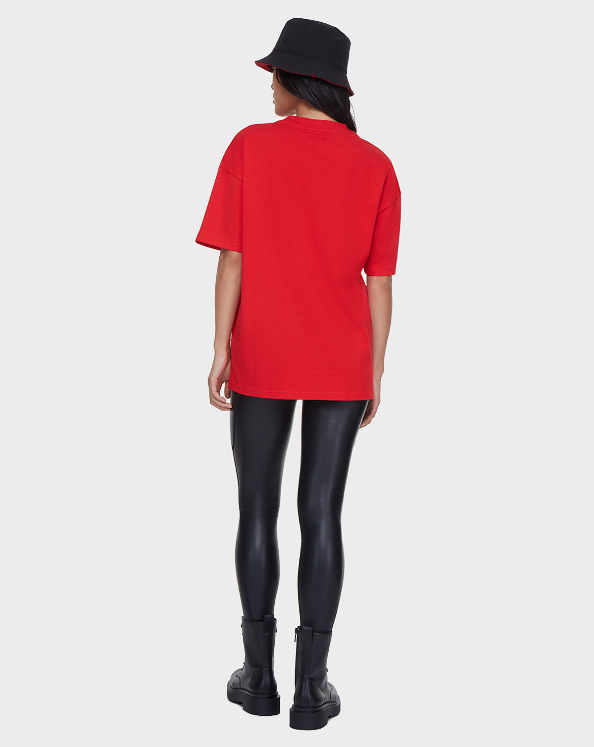 A person stands facing away, wearing the Rudsak LARY - 9521501 POPPY RED oversized unisex t-shirt with the RUDSAK logo, paired with black shiny leggings, black ankle boots, and a black bucket hat against a plain white background.