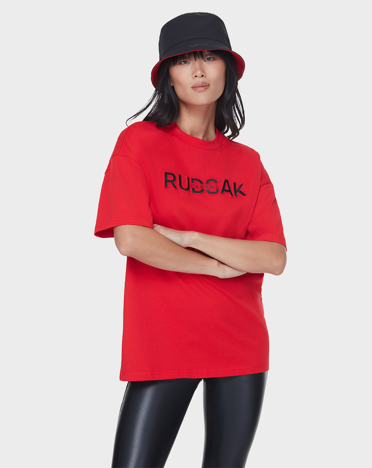 A woman stands with arms crossed against a white background, wearing a black bucket hat, Rudsak's LARY - 9521501 POPPY RED t-shirt with an embossed logo, and black shiny leather-look leggings.