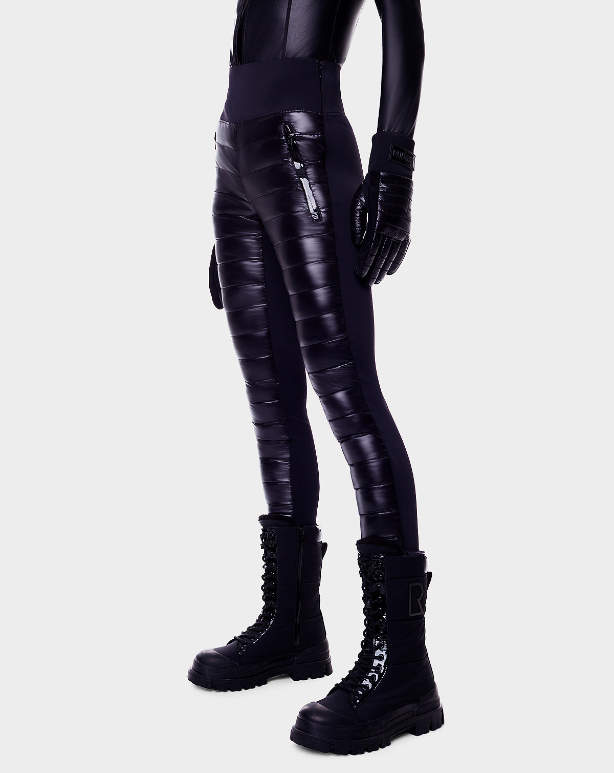 A person is wearing Rudsaks ARIL - 8522520 BLACK shiny quilted padded pants, black gloves, and lace-up boots while standing against a white background. The upper body is cropped out of the image.