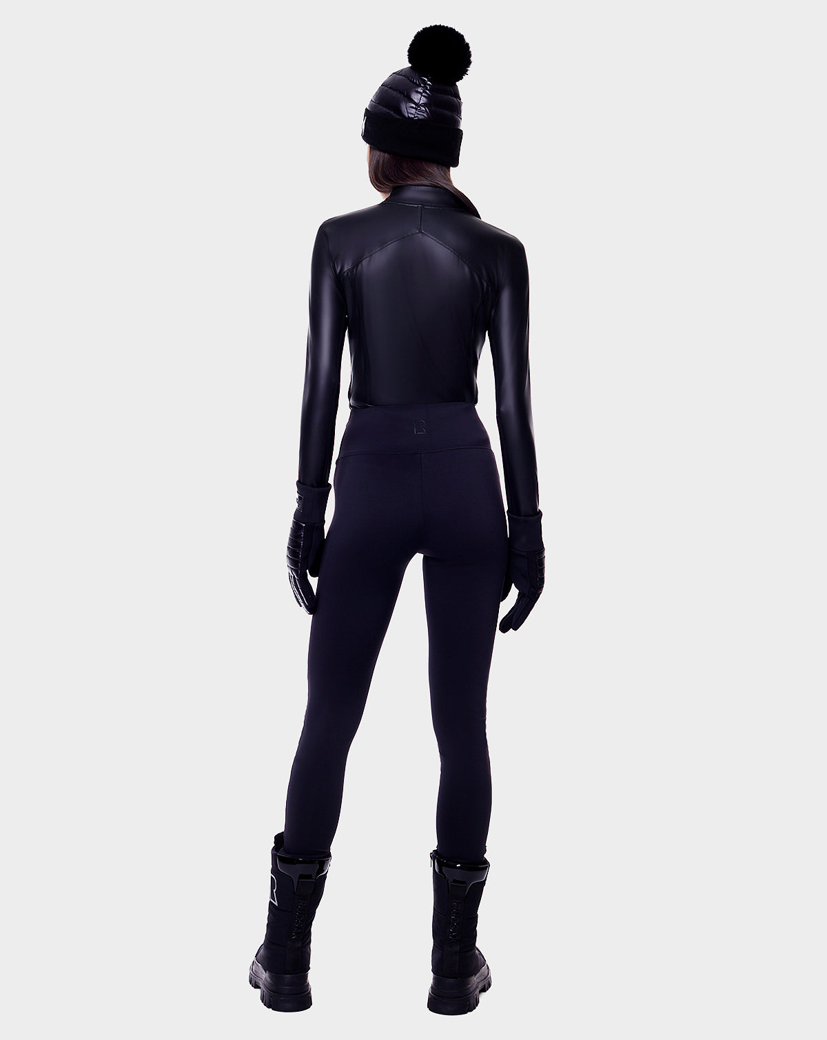 A person faces away, wearing the Rudsak ARIL - 8522520 BLACK: a black fitted jacket with sustainable Sorona fill, paired with a black beanie and gloves, women’s pants, and boots—ideal for an après-ski look against a light gray background.