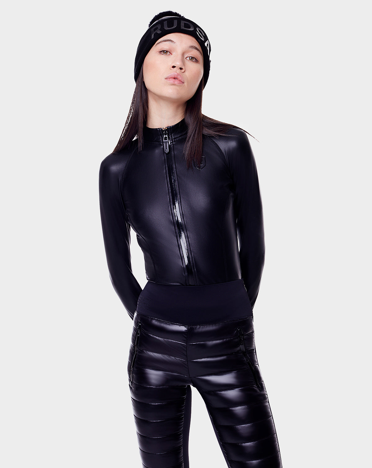 A woman stands against a white background wearing the Rudsak ARIL - 8522520 BLACK beanie, a shiny black zip-up jacket with sustainable Sorona fill, and matching pants—an ideal après-ski look.