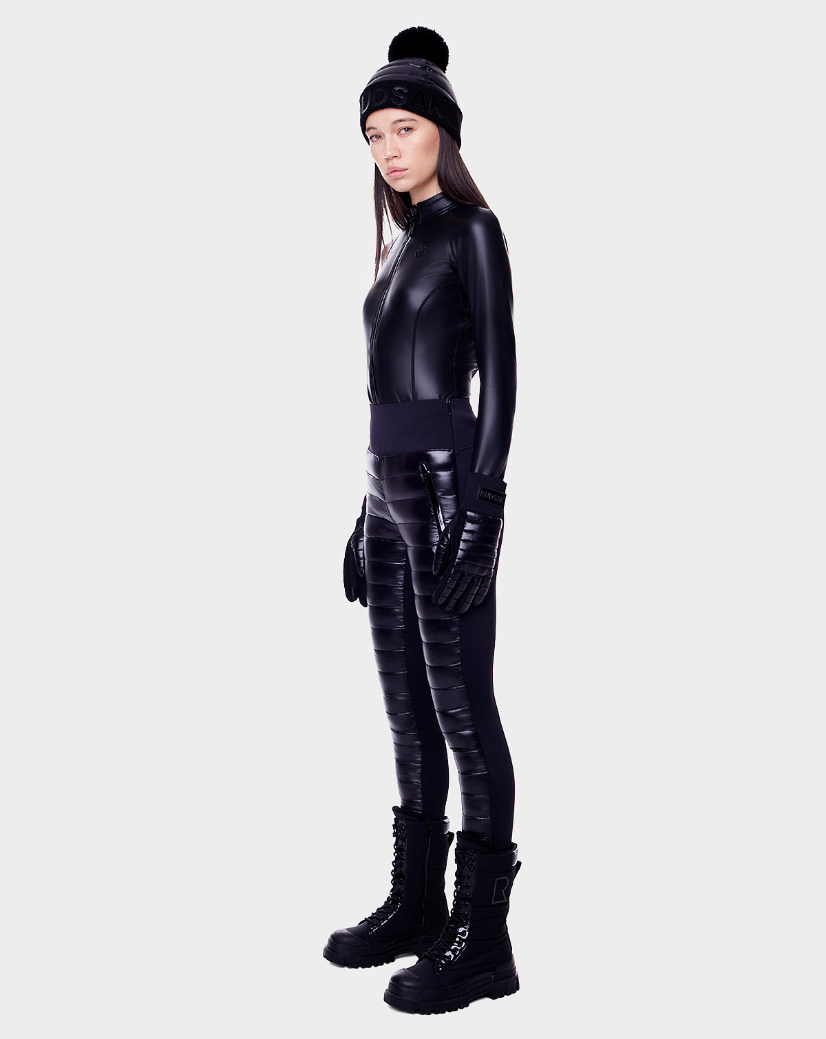 A woman stands against a plain background, wearing the Rudsak ARIL - 8522520 BLACK quilted womens pant with sustainable Sorona fill, paired with a black beanie, gloves, and a fitted leather-like jacket—perfect for après-ski style.