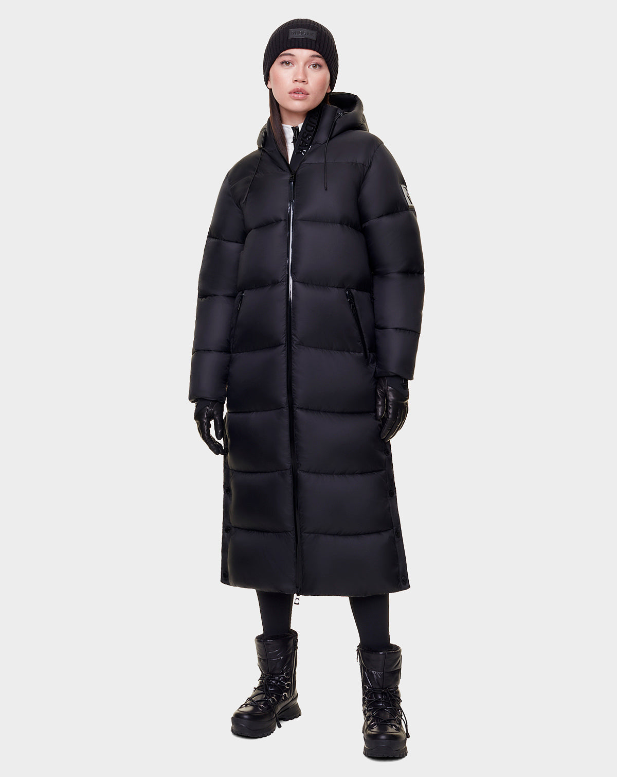 A person wearing the Rudsak EVITA - L122518 BLACK women's down puffer with a hood, black gloves, boots, leggings, and beanie stands against a plain light gray background. The maxi-length coat features waterproof zippers.