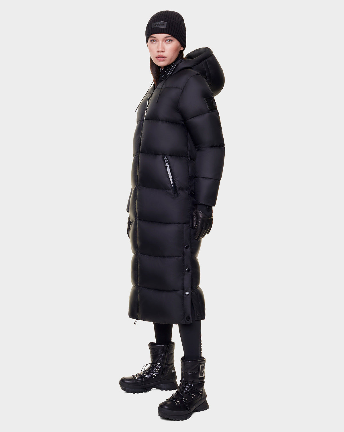 A person wearing the Rudsak EVITA - L122518 BLACK women's down puffer maxi coat with a hood, paired with a black beanie, gloves, leggings, and black lace-up winter boots stands against a plain light background.