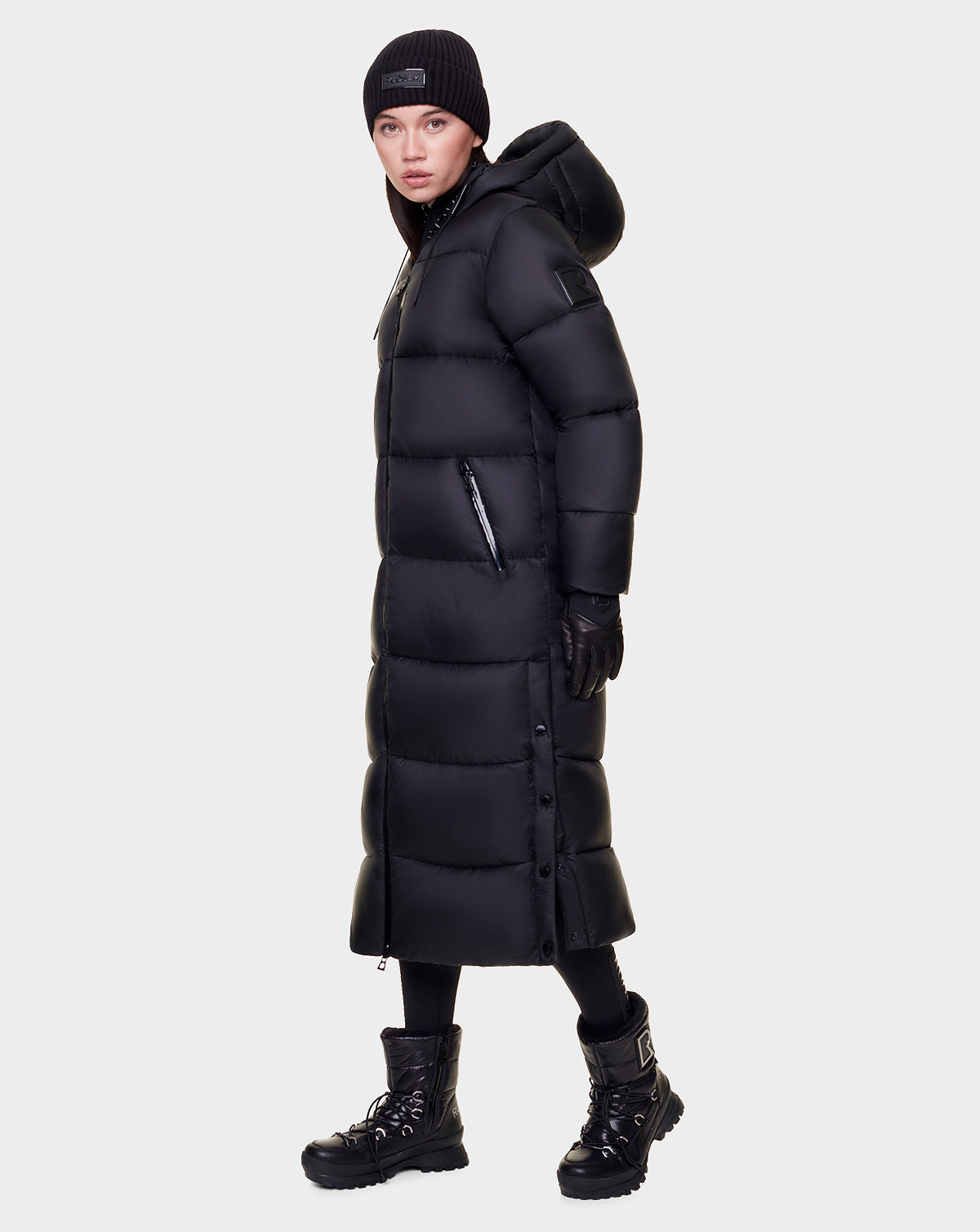 A person models the Rudsak EVITA - L122518 BLACK women's down puffer maxi coat with waterproof zippers, styled with a black beanie, gloves, leggings, and boots, standing against a plain white background and looking slightly to the side.