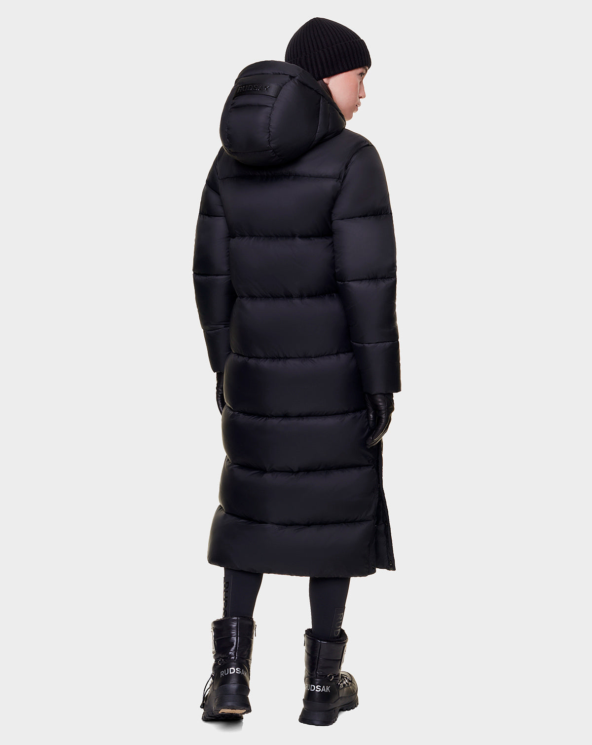 A person wearing the Rudsak EVITA - L122518 BLACK women's down puffer with waterproof zippers, plus a black beanie, gloves, and boots stands facing away against a plain light background.