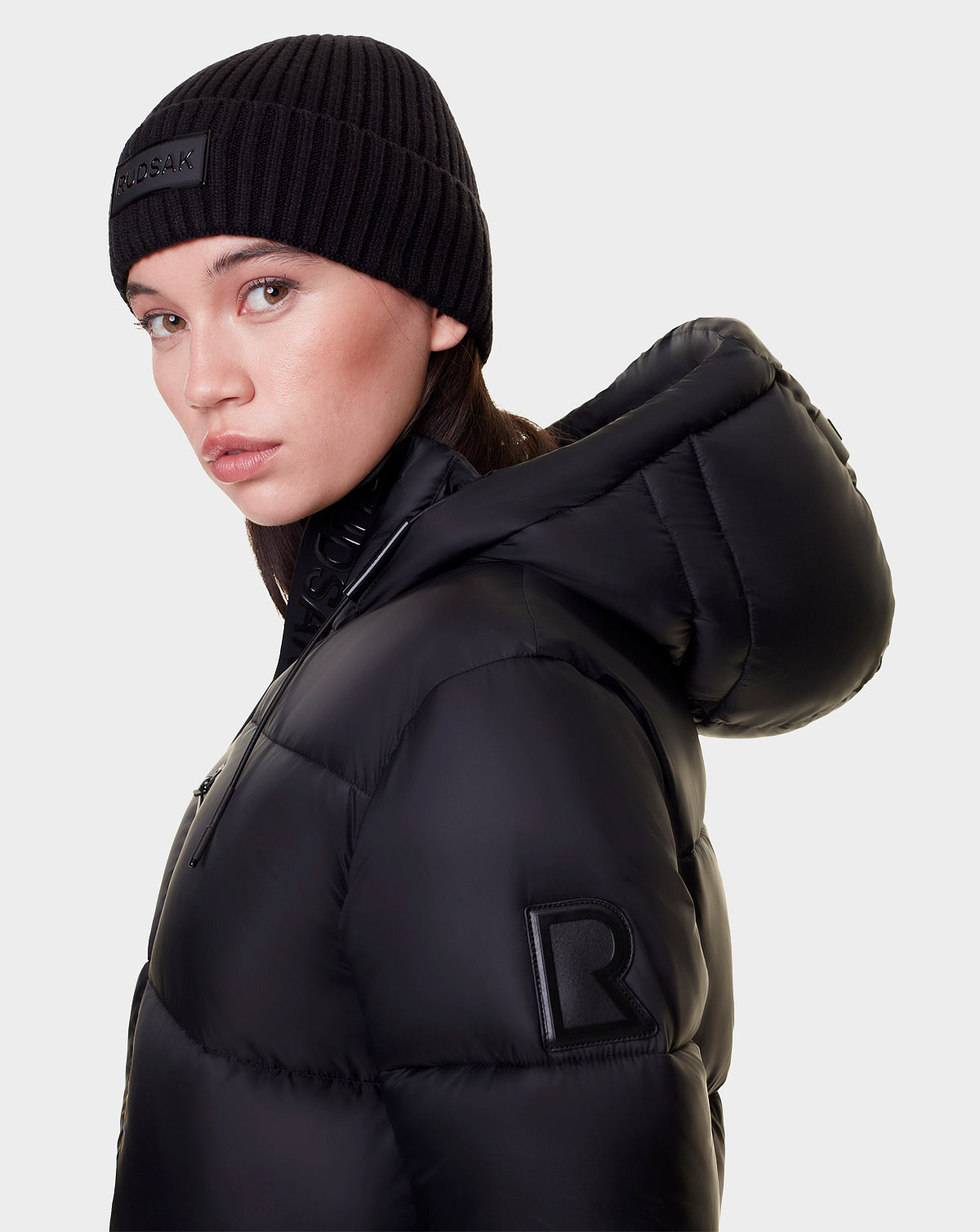 A woman wears the Rudsak EVITA - L122518 BLACK women's down puffer with a large hood and waterproof zippers, plus a black knit beanie. She looks over her shoulder against a plain light background, with an "R" patch on her jacket sleeve.