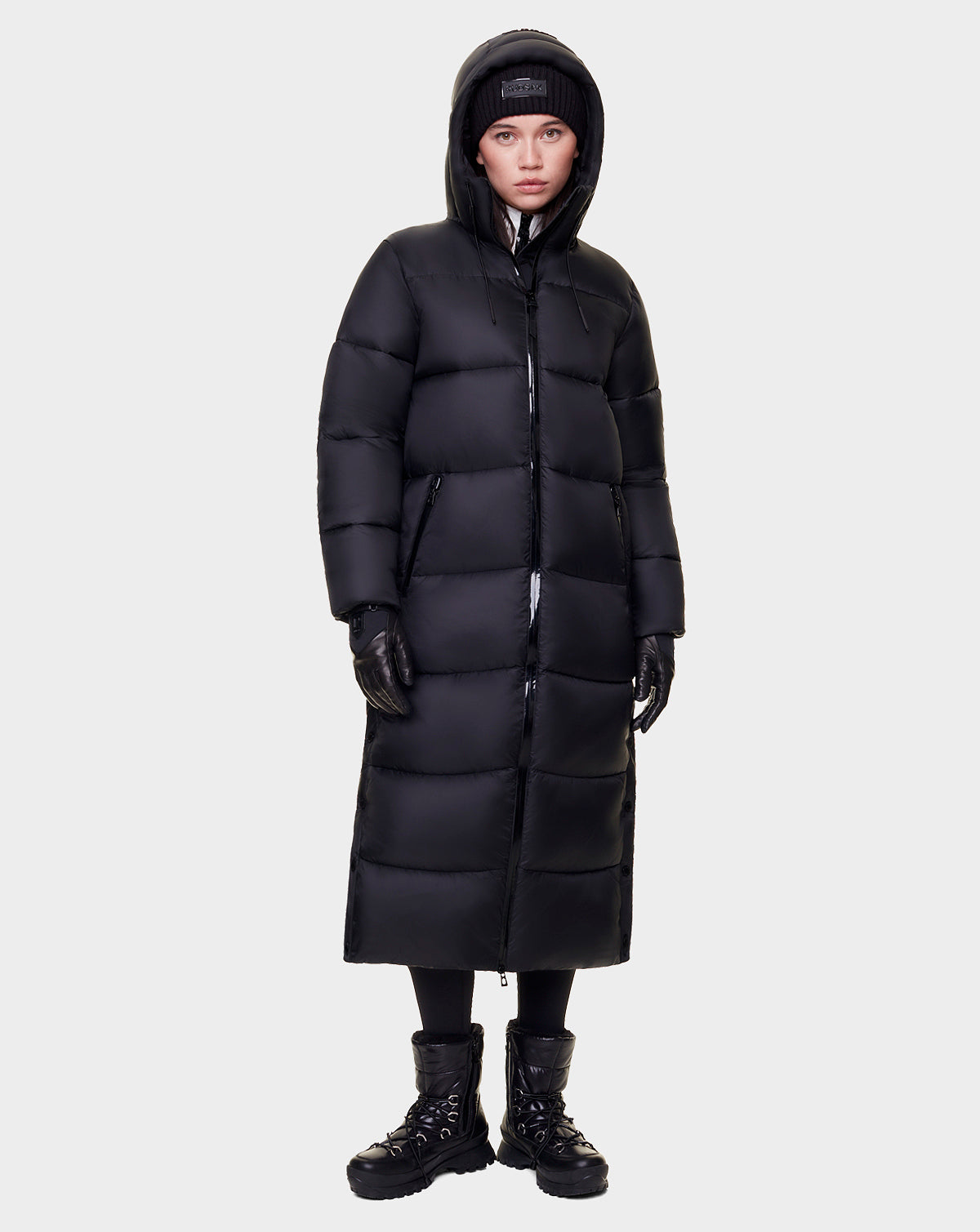 A person stands against a plain background wearing Rudsak's EVITA - L122518 BLACK women's down puffer maxi-length coat with a hood, paired with black gloves, boots, beanie, and pants—a warm winter outfit.