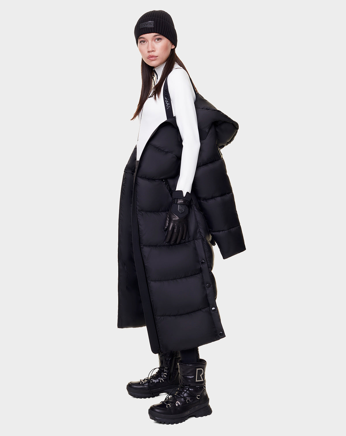 A woman stands sideways wearing the Rudsak EVITA - L122518 BLACK maxi-length down puffer draped over her shoulders, paired with black gloves, boots, a beanie, and a white top. The coat features waterproof zippers, set against a plain light background.