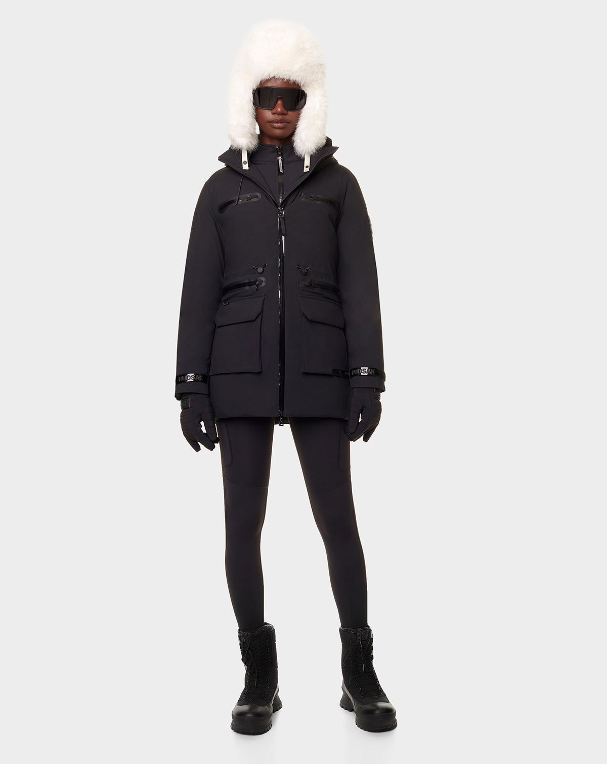 A person wearing the RUDSAK CARLY - 8122514 BLACK down jacket with large front pockets, waterproof zippers, and a white fur hood stands against a plain light gray background.