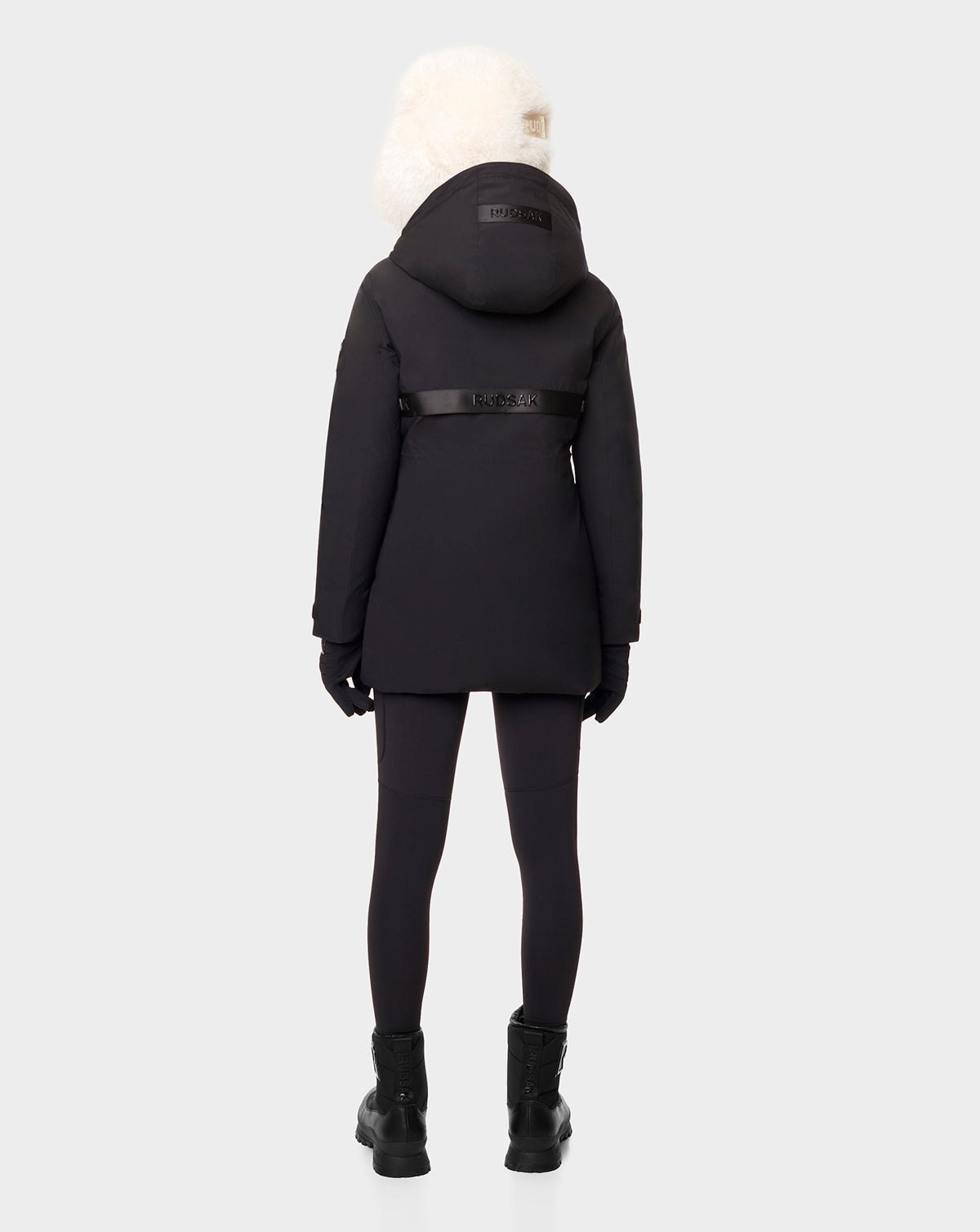 Person stands with their back to the camera wearing the RUDSAK CARLY - 8122514 BLACK hooded down jacket, featuring white fur trim, black leggings, gloves, and black boots. The coat includes RDS-certified down and waterproof zippers. Light gray background.
