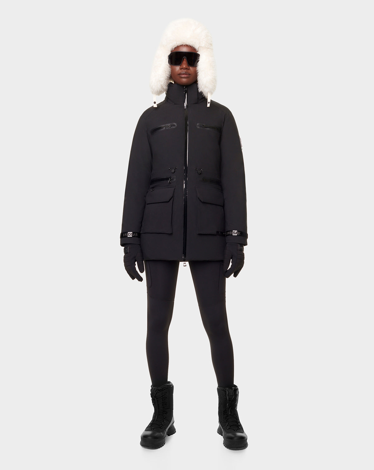 A person wears the RUDSAK CARLY - 8122514 BLACK winter down jacket with waterproof zippers, black leggings, boots, gloves, dark sunglasses, and a white fur-trimmed hood against a plain light gray background.