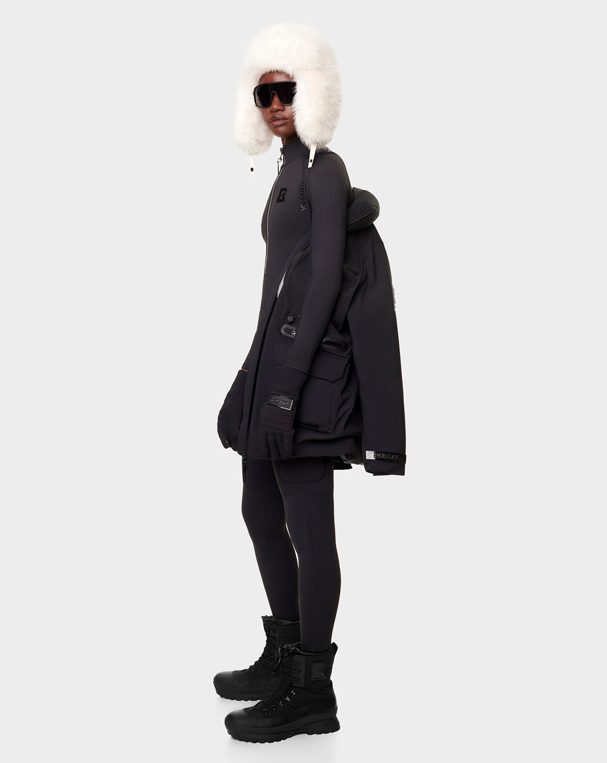 Wearing the RUDSAK CARLY - 8122514 BLACK fitted outfit with black gloves and boots, a large down jacket over one arm, large dark sunglasses, and a fluffy white earflap hat—standing against a light gray background.