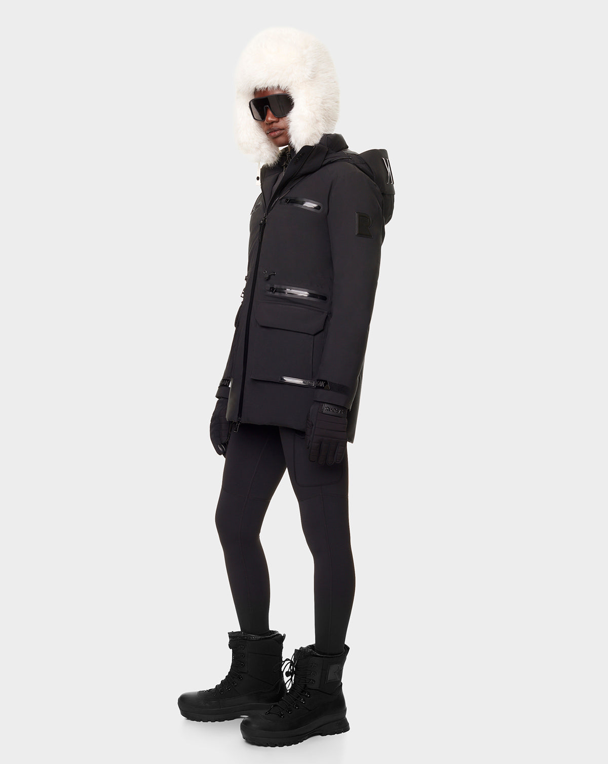 A person wearing the CARLY - 8122514 BLACK down jacket by RUDSAK, with a large white hood, black leggings, gloves, snow boots, and dark sunglasses stands against a plain light background.