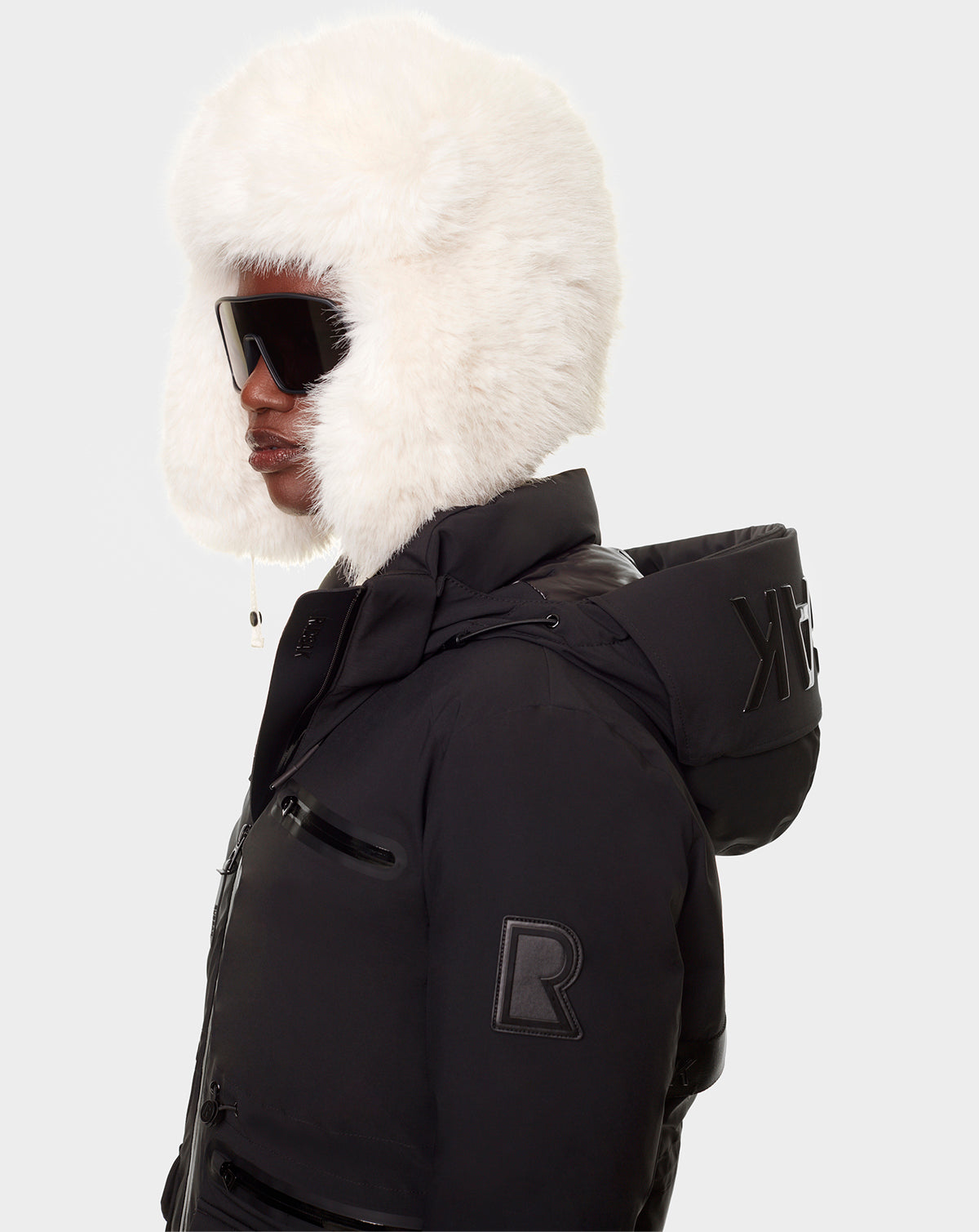 A person wears the RUDSAK CARLY - 8122514 BLACK down parka with waterproof zippers and R & K letters on the collar, paired with a large white fur hat and oversized black sunglasses. The background is plain light gray.