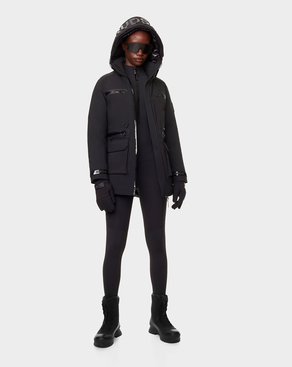 A person wearing the RUDSAK CARLY - 8122514 BLACK winter down jacket, paired with black leggings, gloves, boots, and dark sunglasses stands against a plain light gray background.
