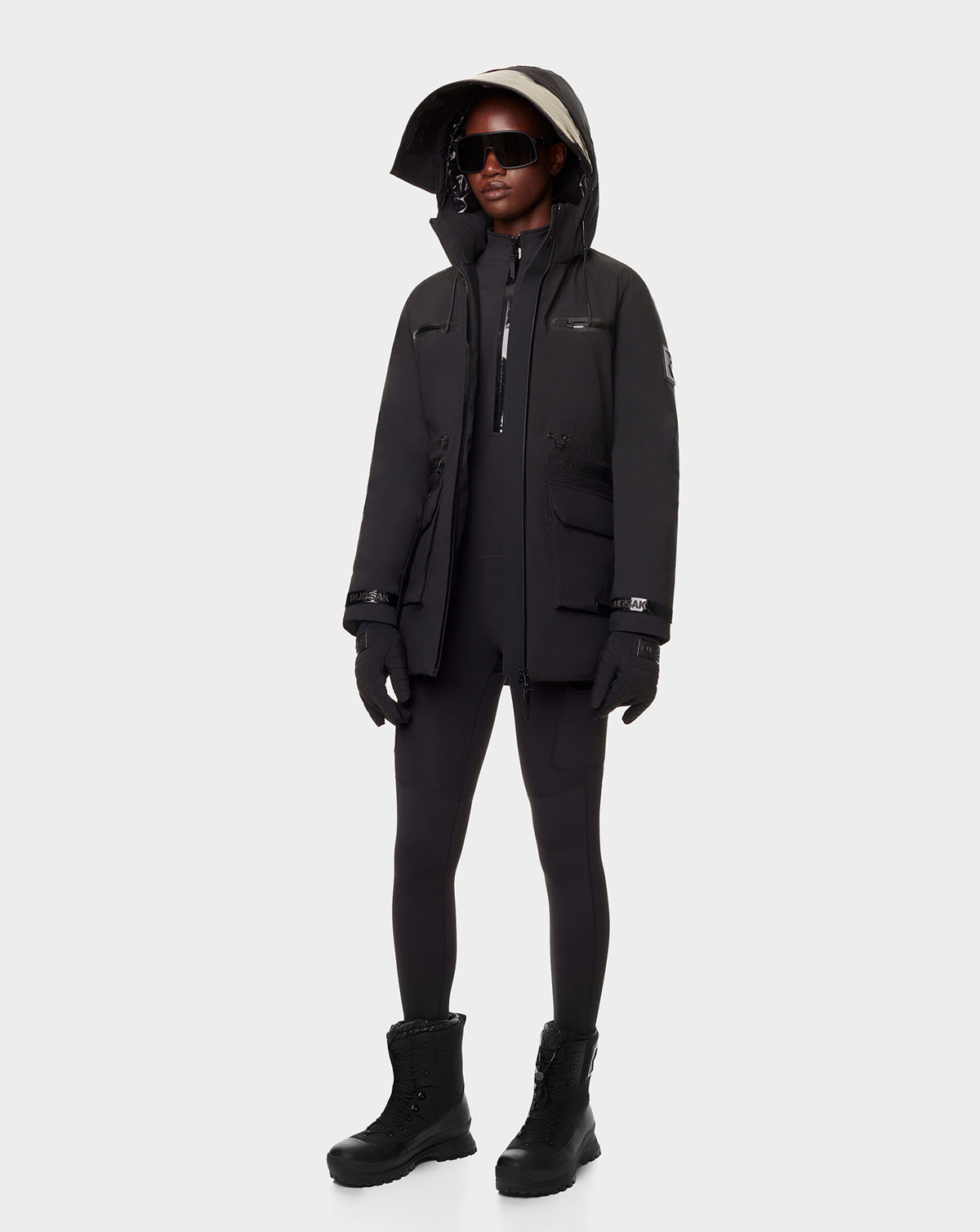 A person wears the RUDSAK CARLY - 8122514 BLACK RDS-certified winter down jacket with waterproof zippers, black leggings, boots, gloves, and dark sunglasses against a plain light background—a warm outfit for cold weather.