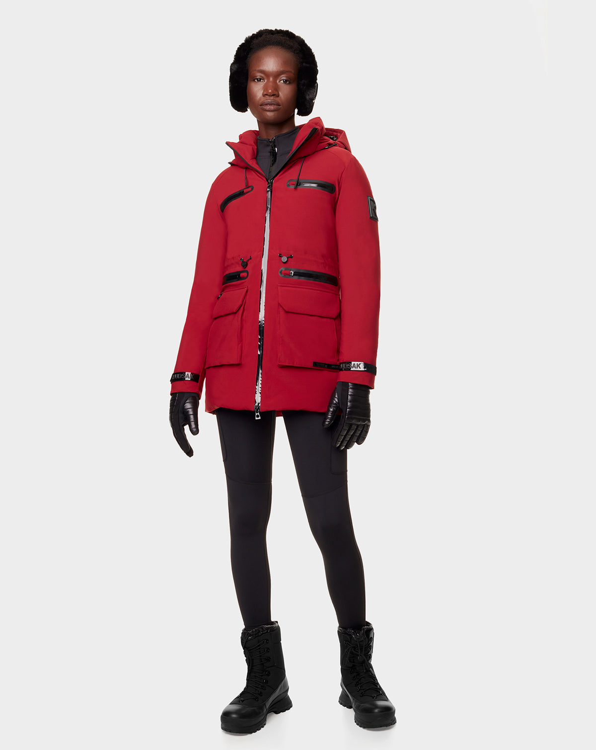 A person wearing the RUDSAK CARLY - 8122514 SANGRIA red hooded winter jacket with waterproof zippers, black gloves, leggings, lace-up boots, and earmuffs stands against a plain light gray background.
