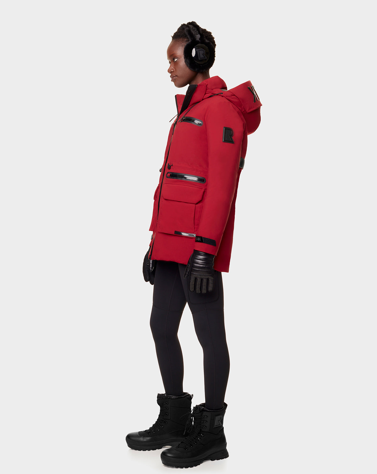 A person in profile wears the RUDSAK CARLY - 8122514 SANGRIA red winter jacket with waterproof zippers, paired with black gloves, leggings, winter boots, and earmuffs against a plain light background.