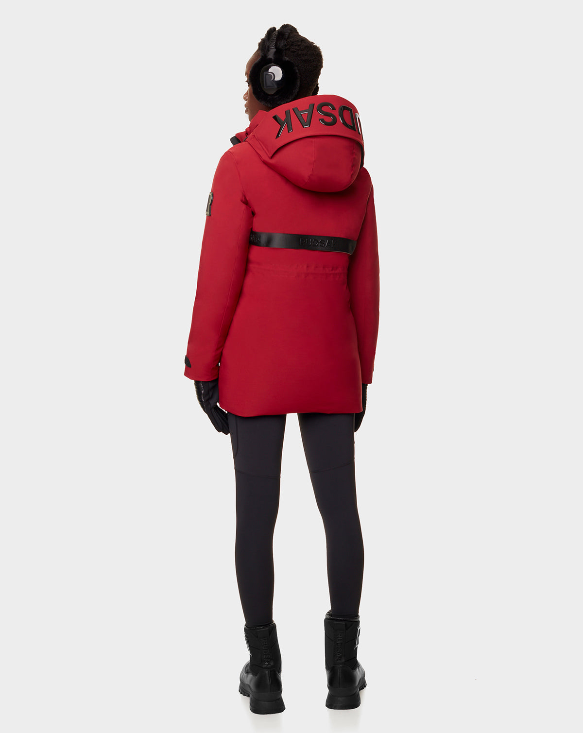 A person is seen from behind wearing the RUDSAK CARLY - 8122514 SANGRIA red winter jacket with a hood featuring large black text and waterproof zippers, paired with black leggings, boots, gloves, and earmuffs.