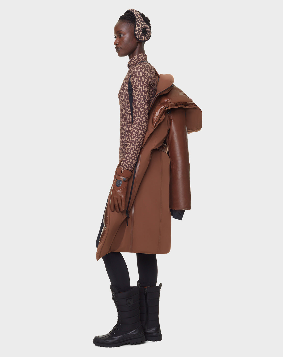 A woman in profile against a white background wears the RUDSAK JANIS - 8122571 COHIBA women's winter coat draped over her shoulder, with a patterned brown top, gloves, black leggings, boots, and a matching headband.