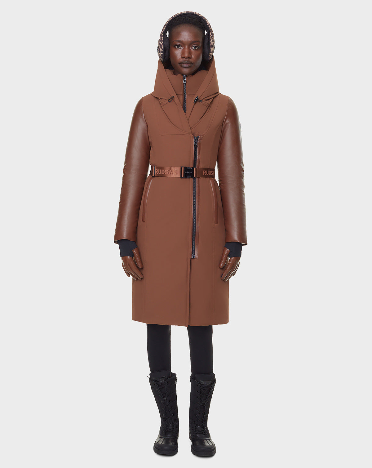 A person wearing the RUDSAK JANIS - 8122571 COHIBA brown belted long down parka with leather sleeves stands facing forward against a plain white background, paired with black gloves, pants, lace-up boots, and earmuffs.