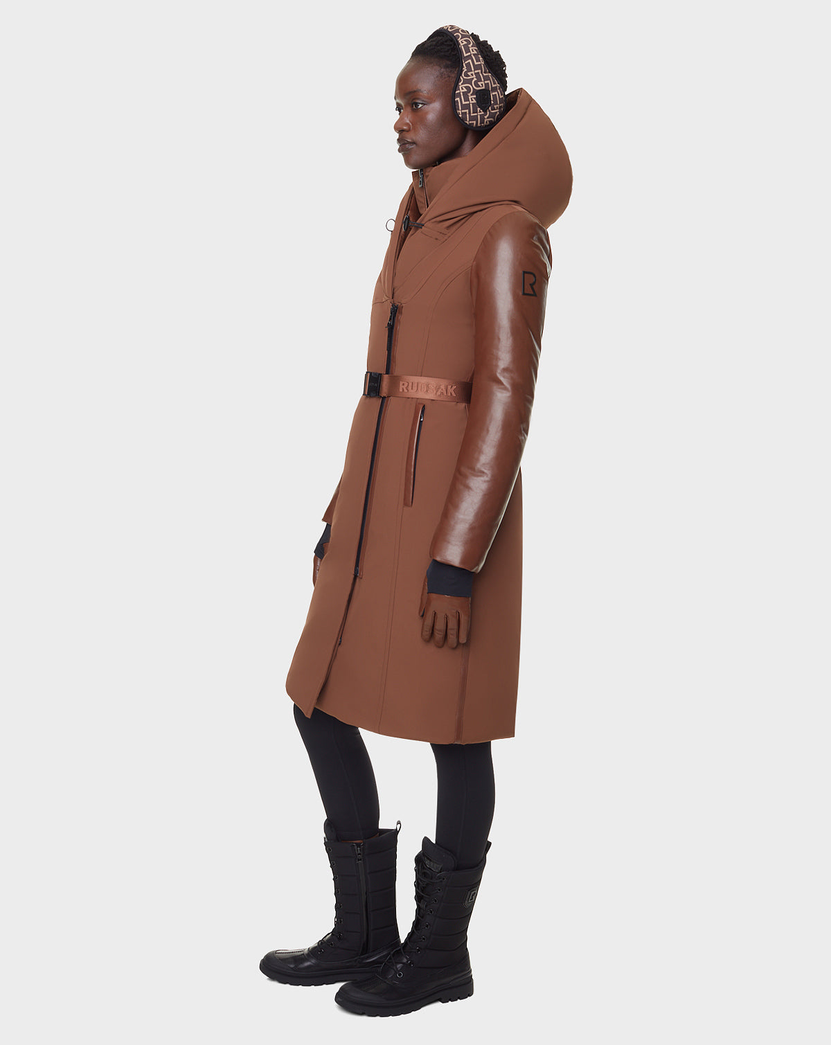 A woman stands in profile against a plain white background wearing the RUDSAK JANIS - 8122571 COHIBA brown belted hooded women’s winter coat, styled with black boots, black leggings, and a patterned headscarf.