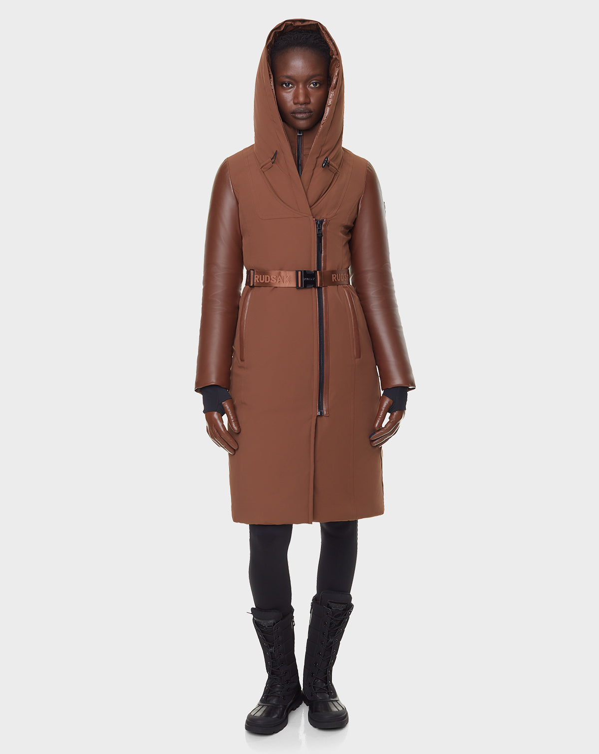 A person faces forward in the RUDSAK JANIS - 8122571 COHIBA women's winter coat with a belted waist, styled like a long down parka, paired with black gloves, leggings, and lace-up boots against a plain white background.