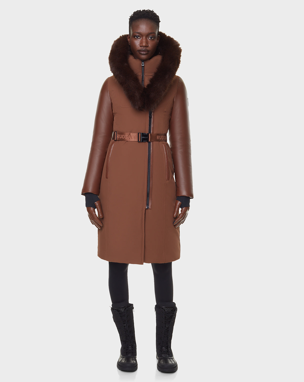 A person stands facing forward, wearing the RUDSAK JANIS - 8122571 COHIBA women’s brown down parka with fur collar and belted waist, paired with black gloves, leggings, and lace-up winter boots against a plain white background.