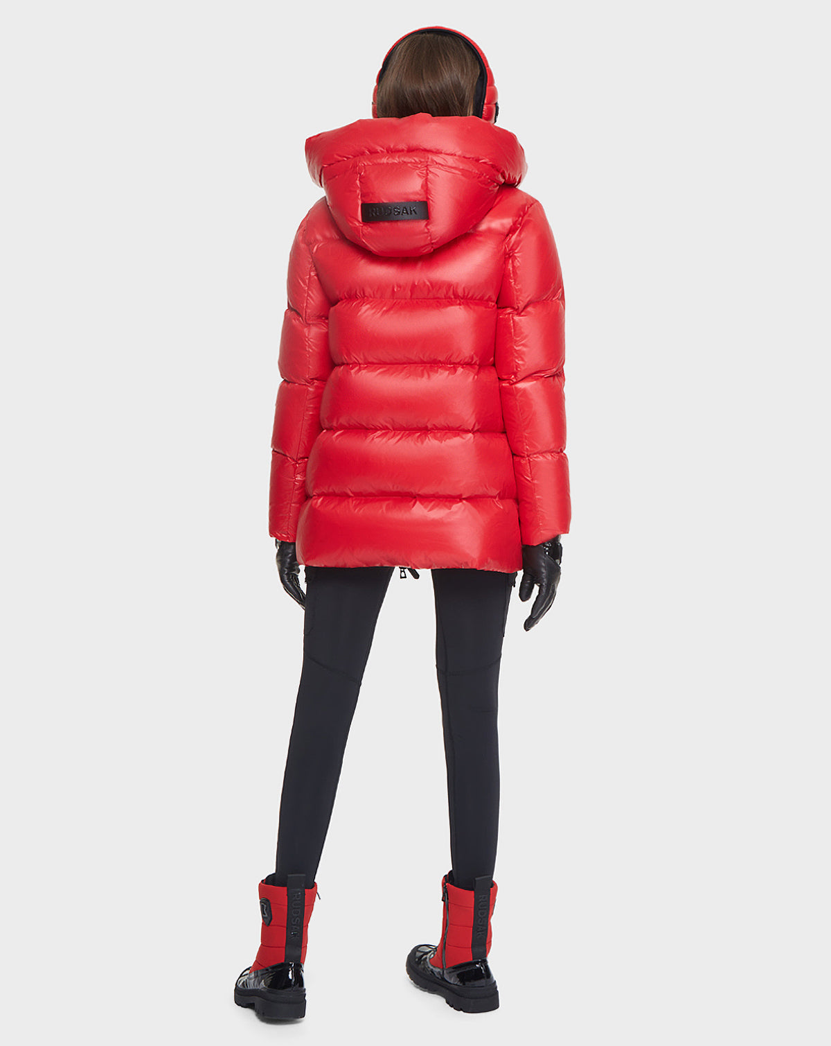 A person faces away, wearing the Rudsak AVALA - L121500 POPPY RED women’s down puffer jacket made from recycled nylon with RDS-certified down, styled with black gloves, fitted pants, red and black boots against a plain white background.