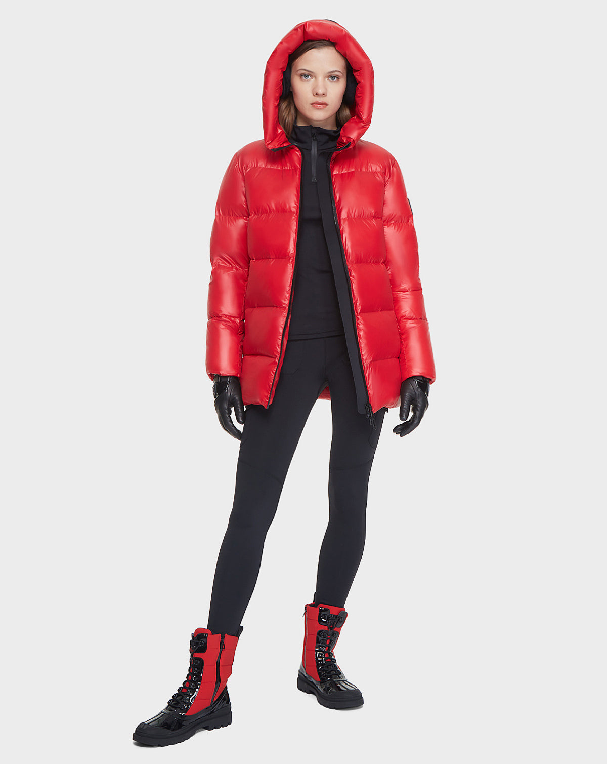 A woman stands facing forward in the Rudsak AVALA - L121500 POPPY RED shiny women's down puffer jacket with hood, crafted from sustainable recycled nylon, paired with black leggings, gloves, and red-and-black lace-up boots against a white background.
