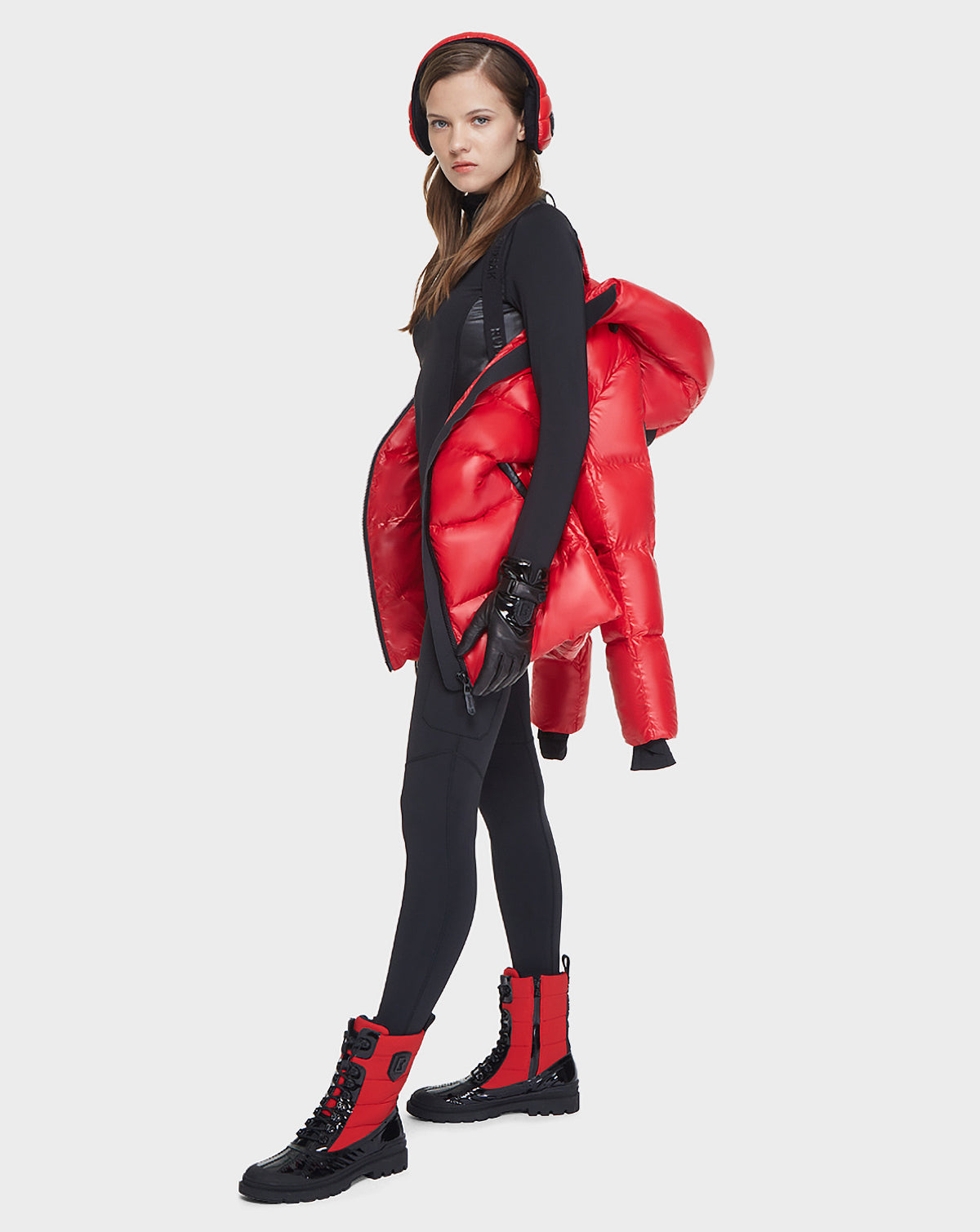 A woman in a black bodysuit stands sideways, looking at the camera against a white background, with the Rudsak AVALA - L121500 POPPY RED women’s down puffer jacket draped over her shoulder and wearing matching red-and-black boots.