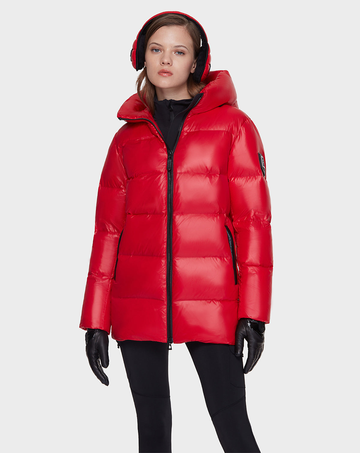 A woman stands facing forward against a white background, wearing the Rudsak AVALA - L121500 POPPY RED down puffer jacket, paired with black gloves, black pants, and red earmuffs.