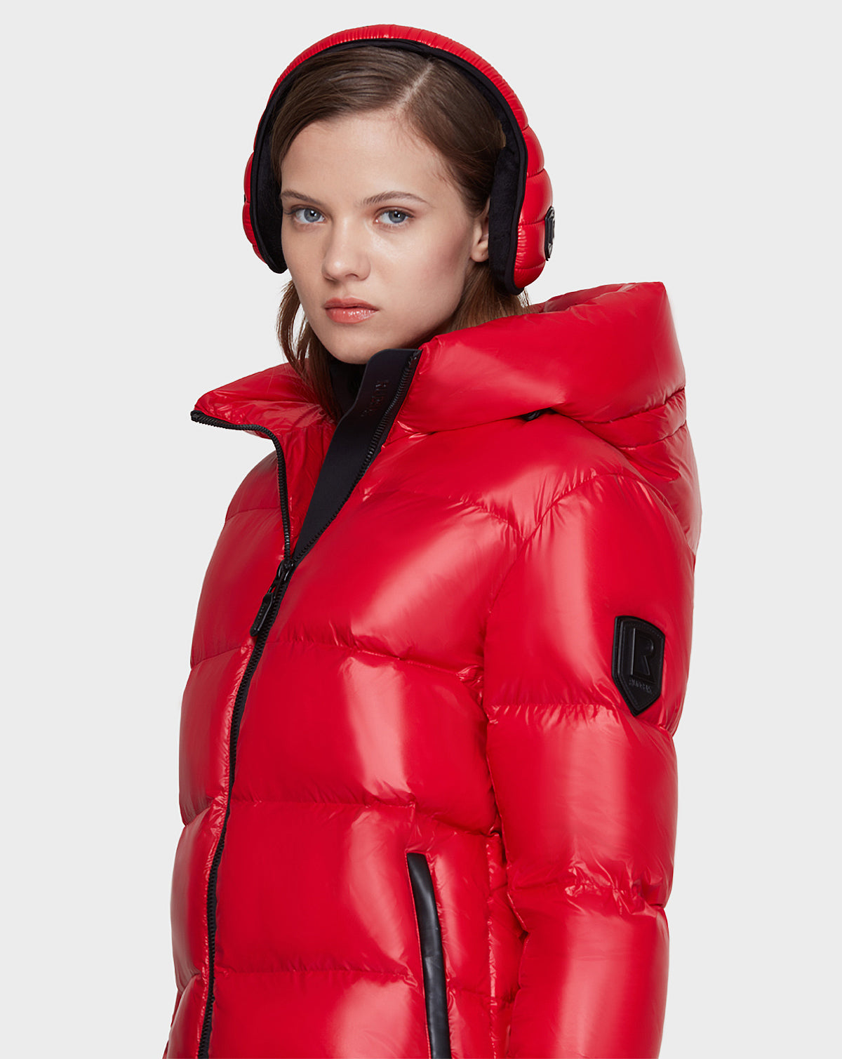 A young woman models the Rudsak AVALA - L121500 POPPY RED women’s down puffer, crafted from recycled nylon with RDS-certified down and black trim, against a light gray background, looking at the camera neutrally.