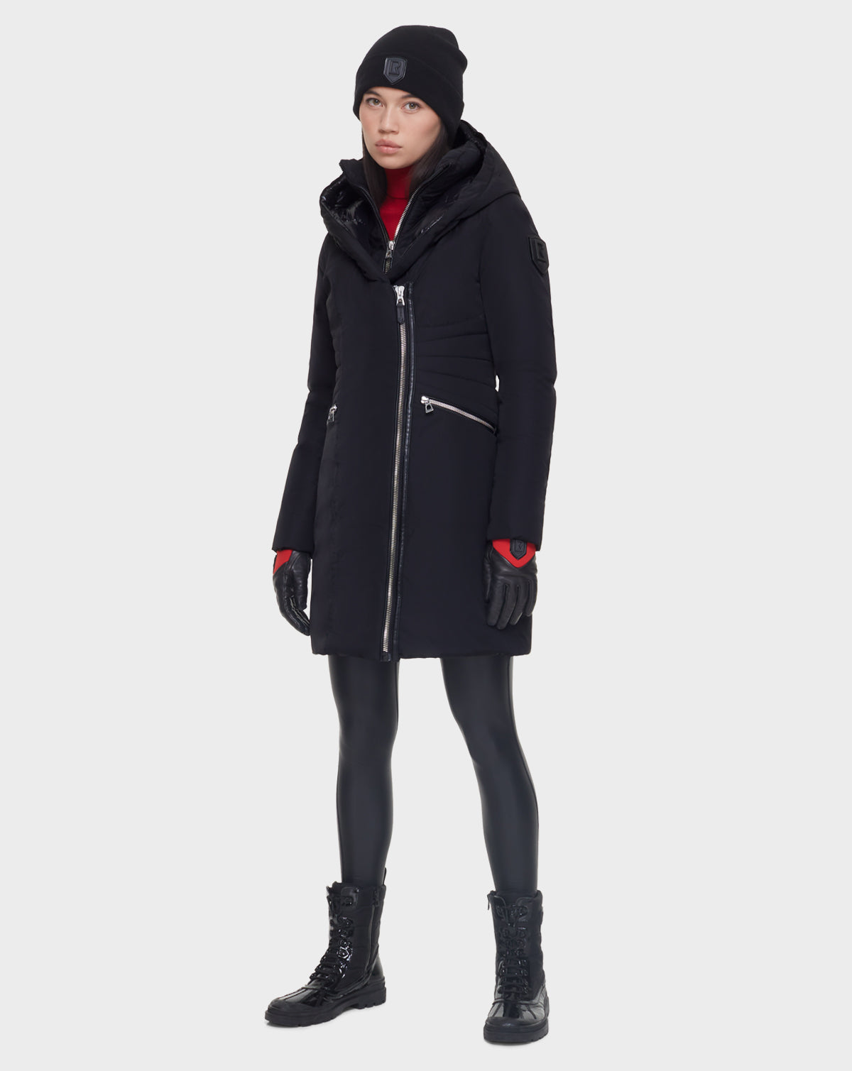A woman faces forward against a plain light background, wearing the Rudsak CORA - L121526 BLACK women’s fitted parka with an asymmetrical zipper, a black beanie, gloves, shiny leggings, and lace-up boots.
